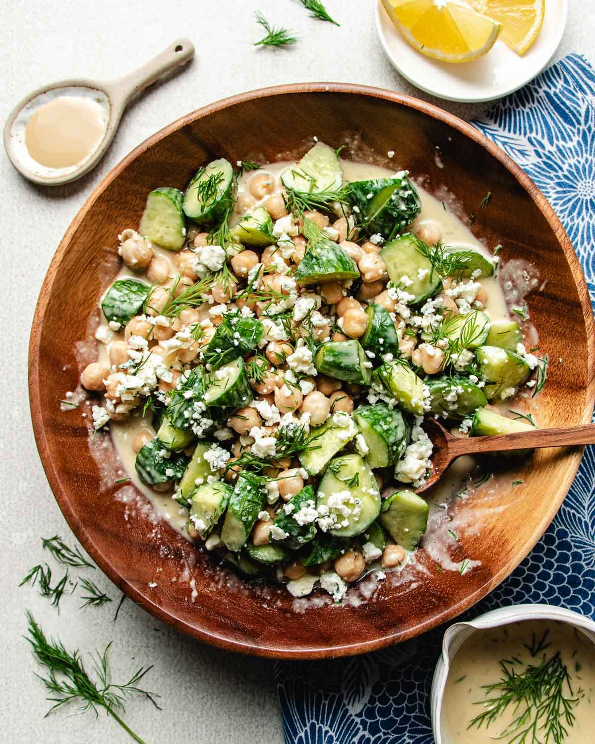 A wooden bowl of cucumber chickpea salad with fresh dill, and crumbled blue cheese sits with a wooden spoon. Nearby are a small bowl of creamy dressing and lemon wedges on a blue patterned cloth.