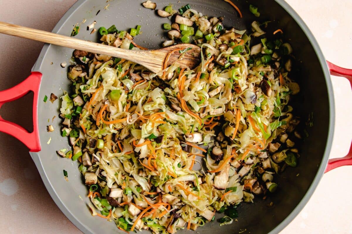 A large skillet with red handles contains sautéed vegetables, including shredded cabbage, carrots, mushrooms, and green onions —perfect dumpling filling ideas for rice paper wrapped dumplings. A wooden spoon rests in the skillet, stirring the colorful mixture.