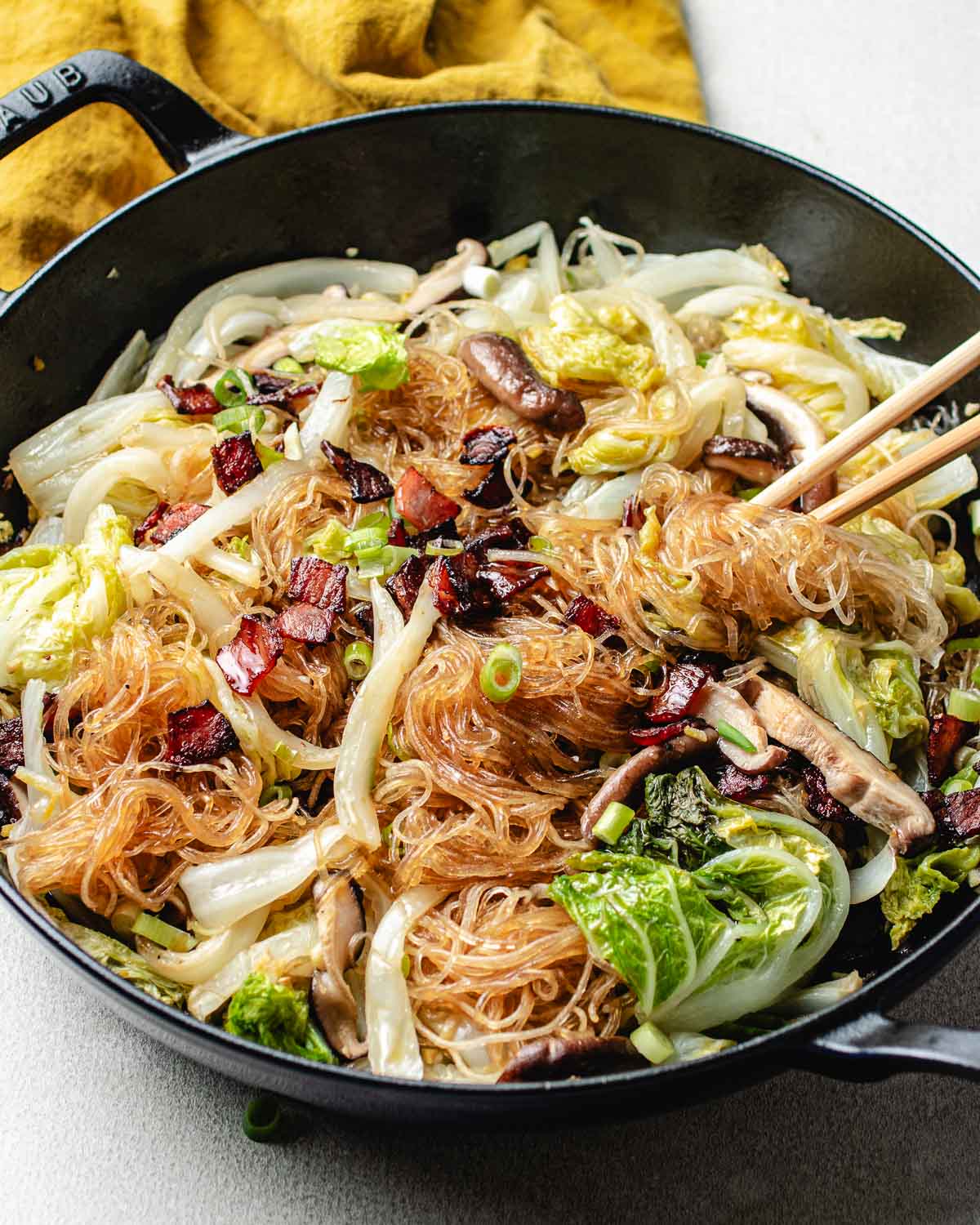A skillet filled with steamed Napa cabbage with glass noodles, mushrooms, and pieces of cooked bacon, with a pair of chopsticks resting on top. A yellow cloth is visible in the background.