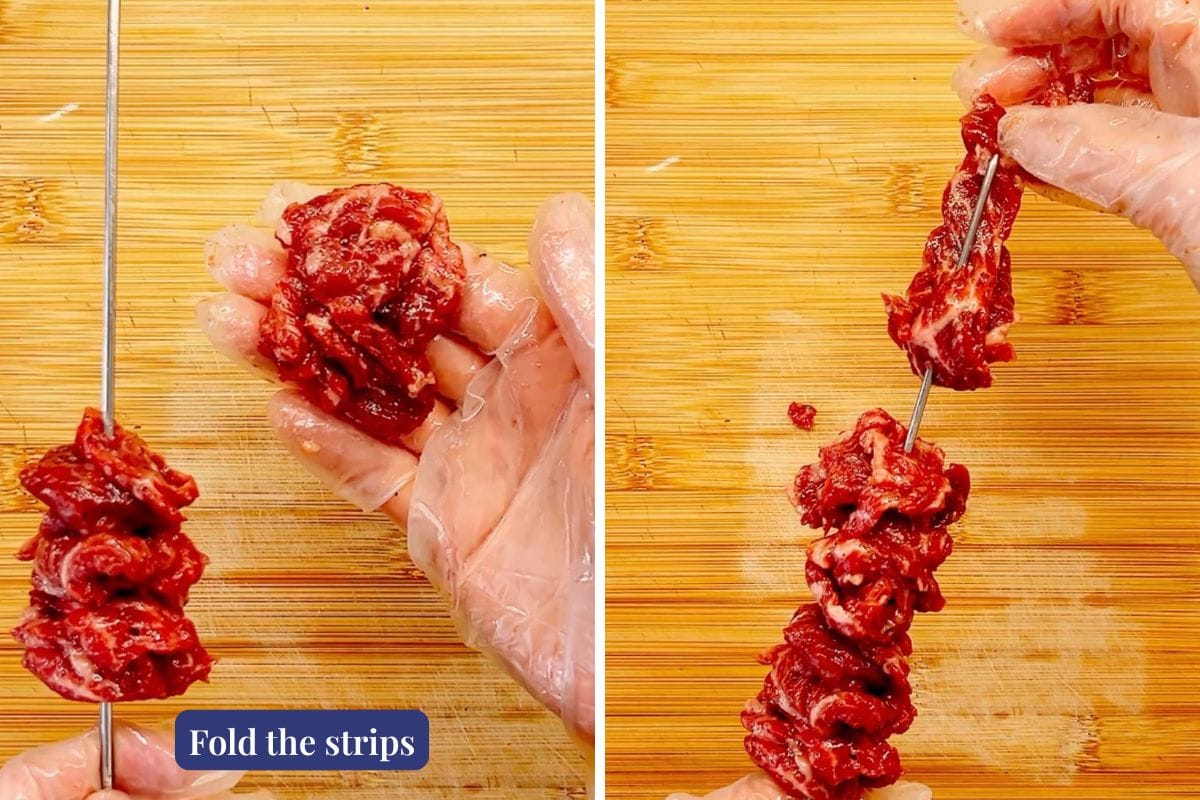 Close-up of two gloved hands threading marinated beef strips onto a metal skewer over a wooden cutting board—perfect for making beef skewers with ginger scallion sauce. A label at bottom left reads, Fold the strips for easy threading.