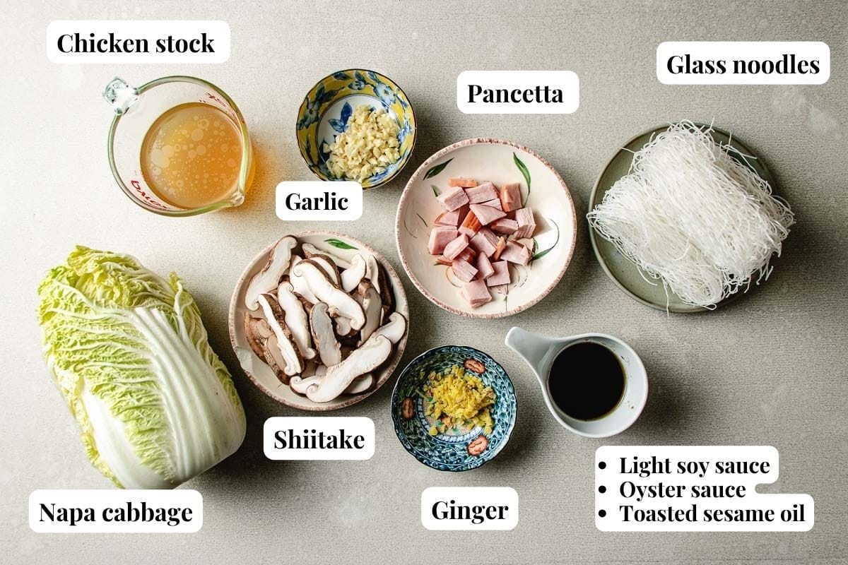 Top-down view of ingredients in bowls for a noodle dish: chicken stock, pancetta, glass noodles, napa cabbage, shiitake mushrooms, garlic, ginger, and sauces (light soy, oyster sauce, toasted sesame oil).