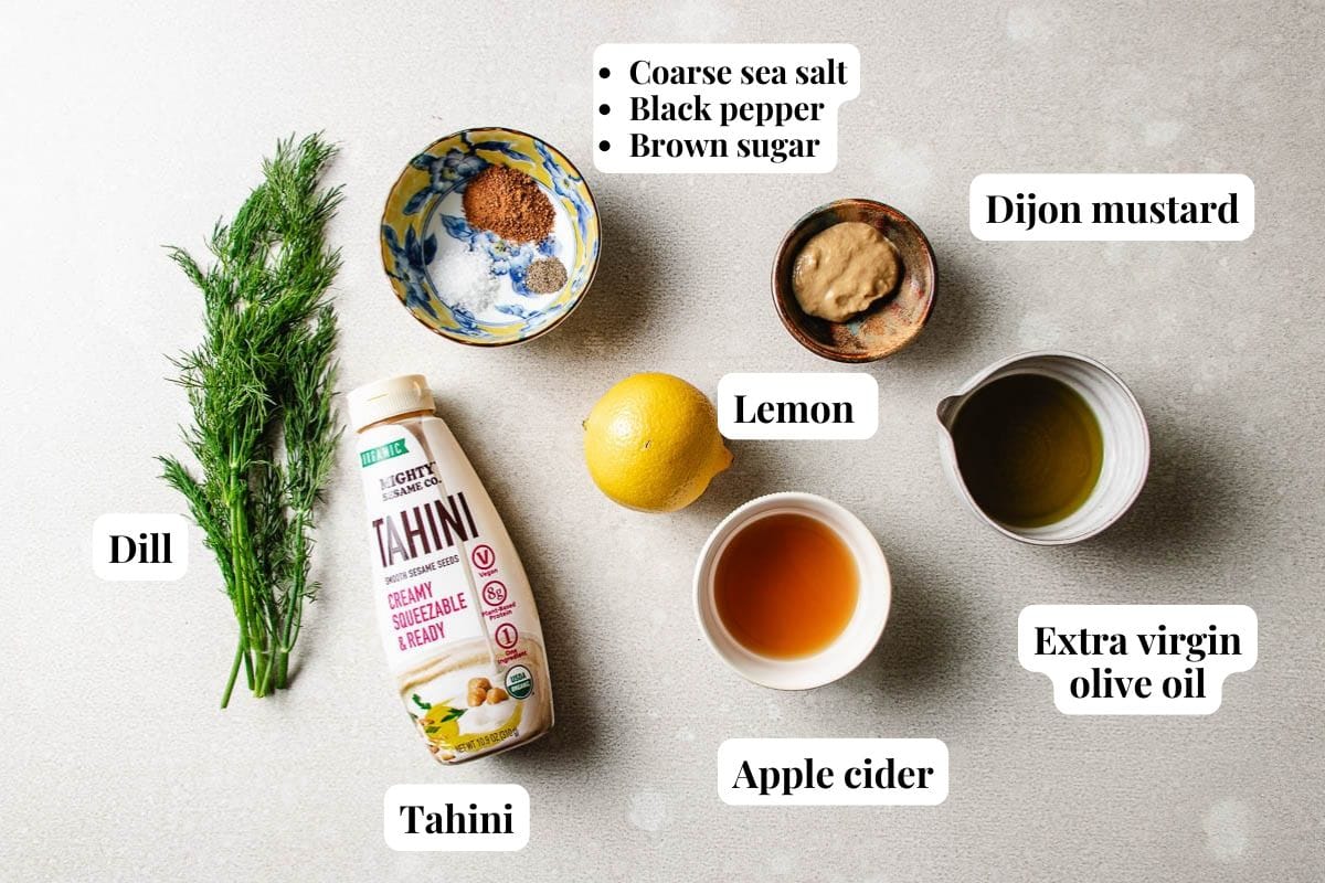Ingredients for a Tahini dill dressing are labeled and laid out on a light surface: tahini, dill, lemon, apple cider, Dijon mustard, extra virgin olive oil, plus coarse sea salt, black pepper, and brown sugar in a bowl.