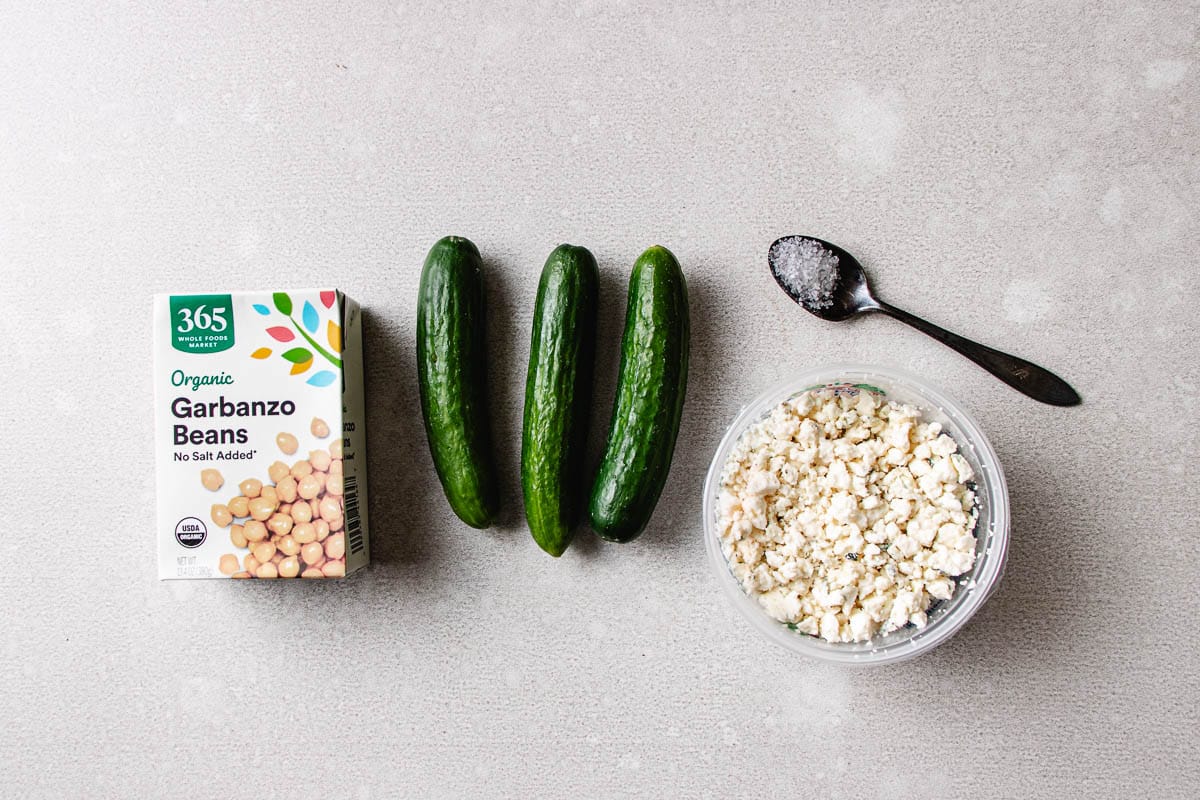 Box of organic garbanzo beans, three cucumbers, crumbled cheese, and a spoon with salt arranged on a light gray surface—perfect for making a fresh cucumber chickpea salad.