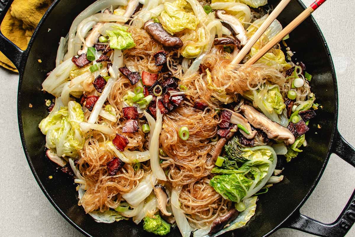 steamed Napa cabbage with cellophane noodles, shiitake mushrooms, scallions, and crispy pancetta pieces in a black skillet. Chopsticks rest on the side, ready for serving.