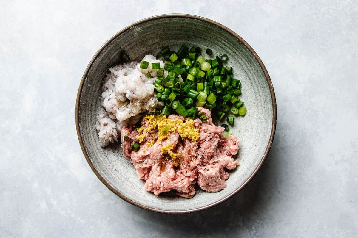 A ceramic bowl containing ground pork and finely minced shrimp, chopped green onions, grated ginger, and toasted sesame oil—all arranged in separate sections—offers fresh dumpling filling ideas for siu mai-style rice paper rolls on a light surface.