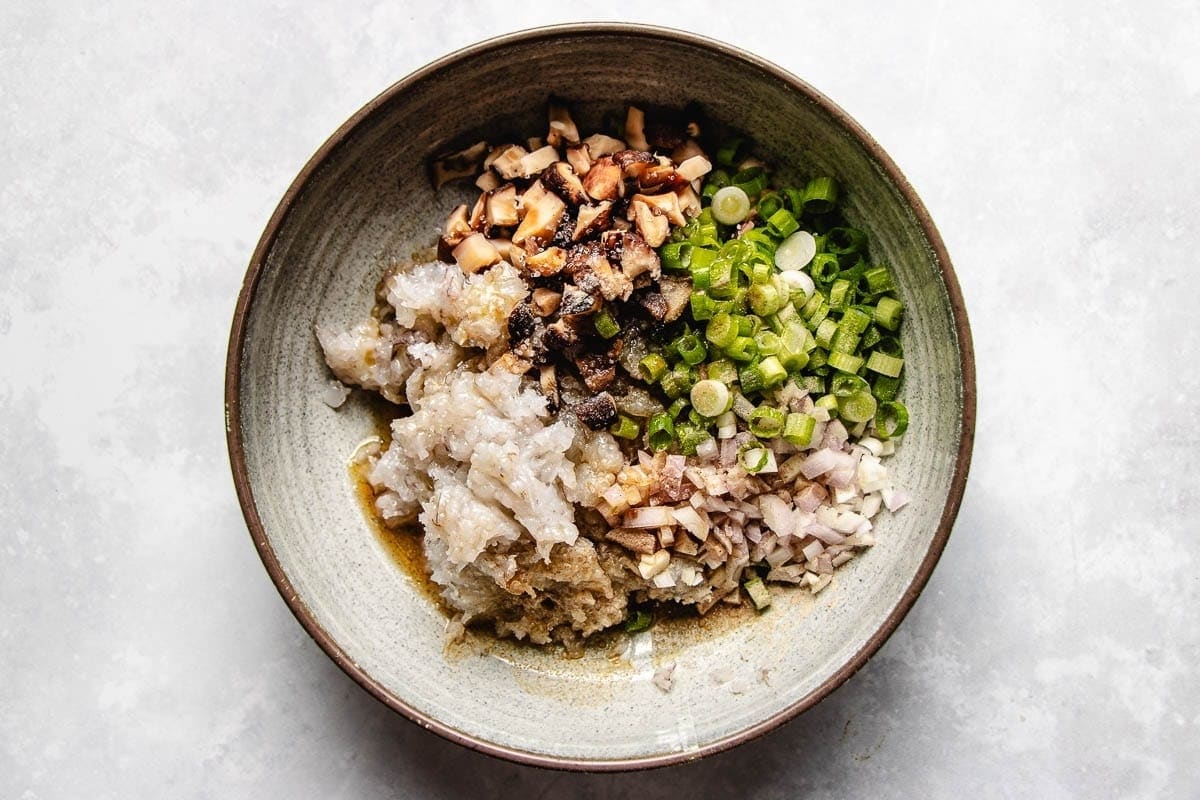 A bowl containing separated piles of chopped green onions, diced mushrooms, and a ground meat mixture—perfect dumpling filling ideas for rice paper—ready to be mixed for a recipe, on a light surface.