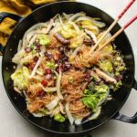 A black skillet filled with steamed Napa cabbage, cellophane noodles, shiitake, crispy bacon pieces, and green onions, with a pair of chopsticks resting on top.