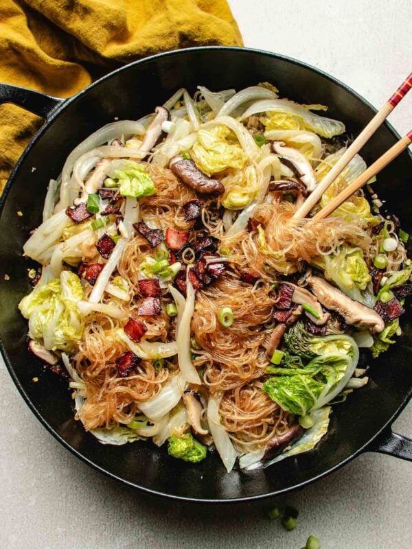 A black pan filled with napa cabbage, bean thread noodles, mushrooms, green onions, and pancetta bits sit beside red chopsticks. Steamed Napa cabbage adds freshness, while a yellow cloth is partially visible in the corner.