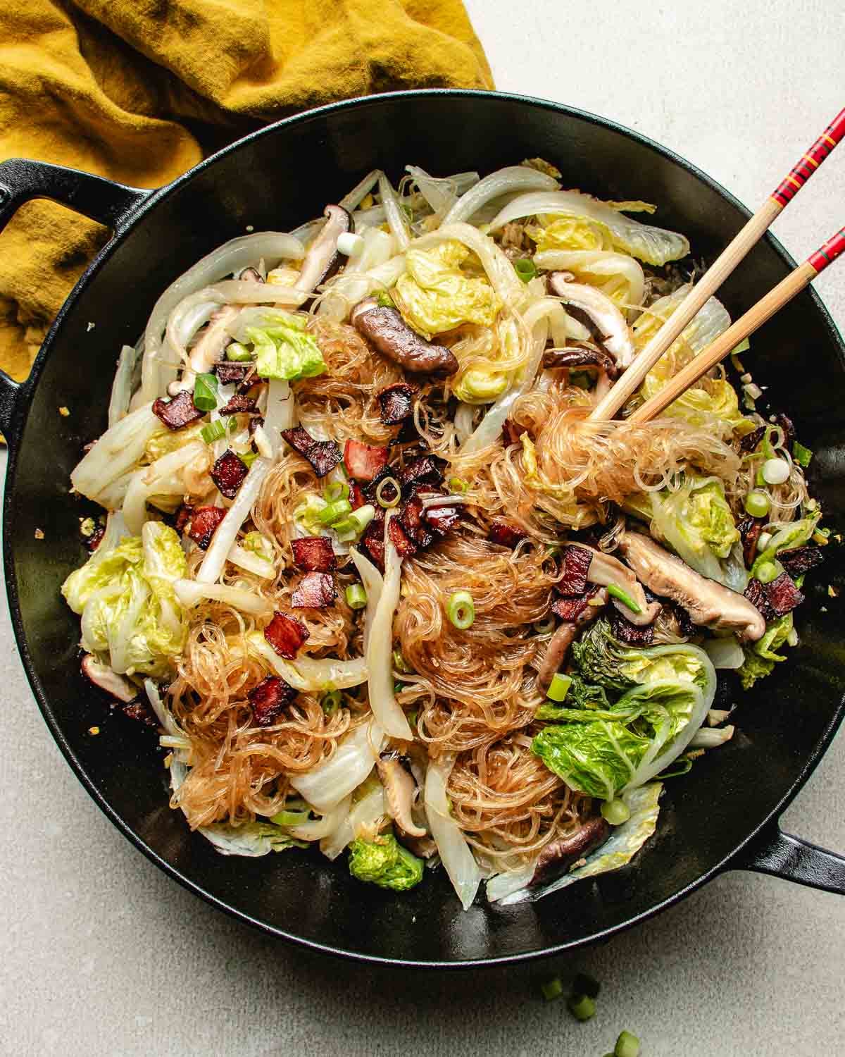 A black pan filled with napa cabbage, bean thread noodles, mushrooms, green onions, and pancetta bits sit beside red chopsticks. Steamed Napa cabbage adds freshness, while a yellow cloth is partially visible in the corner.