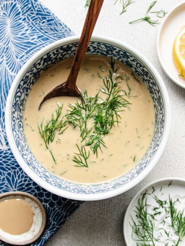 A bowl of creamy tahini dill dressing garnished with fresh dill, with a wooden spoon resting inside. Nearby are a lemon wedge, a small dish of dill, and extra tahini, all on a blue and white patterned cloth.