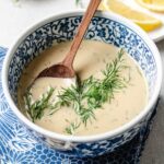 A blue and white patterned bowl filled with creamy tahini lemon dill dressing, garnished with fresh dill, and a wooden spoon resting inside. Slices of lemon are visible on a plate in the background.