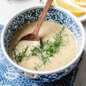A blue and white patterned bowl filled with creamy tahini lemon dill dressing, garnished with fresh dill, and a wooden spoon resting inside. Slices of lemon are visible on a plate in the background.