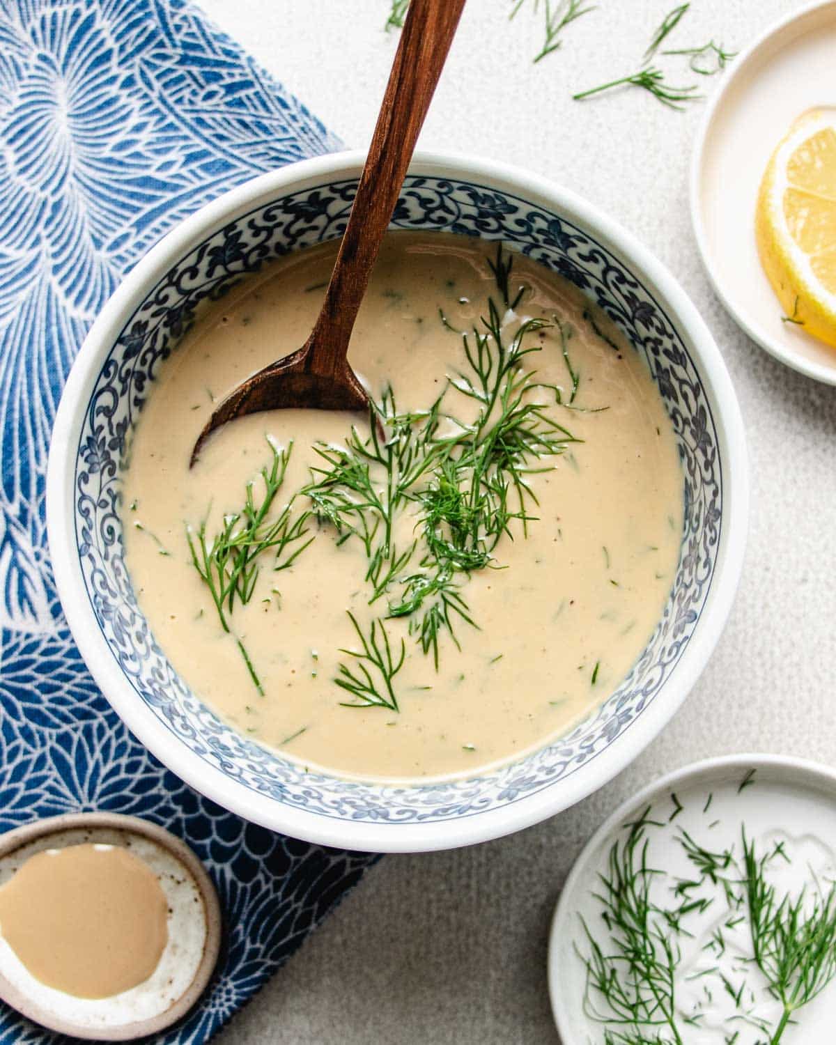 A bowl of creamy tahini dill dressing garnished with fresh dill, with a wooden spoon resting inside. Nearby are a lemon wedge, a small dish of dill, and extra tahini, all on a blue and white patterned cloth.