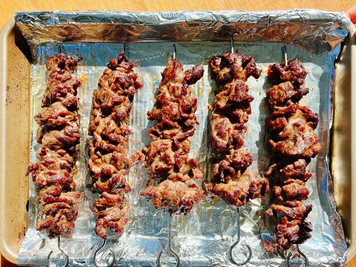 Five metal skewers of grilled beef sit on a foil-lined baking tray, the meat cooked to a little char outside and juicy tender inside, arranged in rows under bright sunlight.