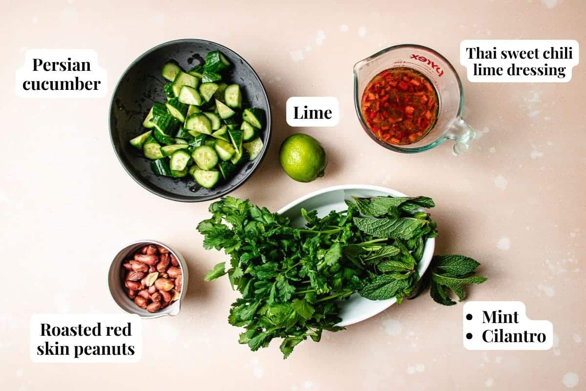 Bowls and containers with chopped Persian cucumber, roasted red skin peanuts, a whole lime, Thai sweet chili lime dressing, fresh mint, and cilantro on a light surface—perfect for assembling your own Thai cucumber shrimp salad. Text labels identify each ingredient.