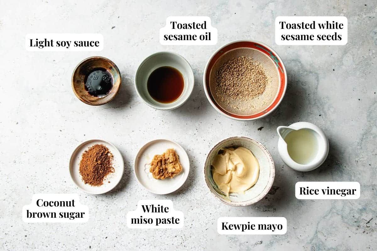 Seven small bowls with labeled ingredients for sesame miso dressing on a light surface: light soy sauce, toasted sesame oil, toasted white sesame seeds, coconut brown sugar, white miso paste, Kewpie mayo, and rice vinegar.