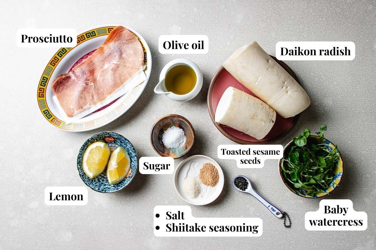 A flat lay of ingredients for a shaved daikon salad—prosciutto, olive oil, baby watercress, toasted sesame seeds, shiitake seasoning, salt, sugar, and lemon quarters—arranged on a light surface.