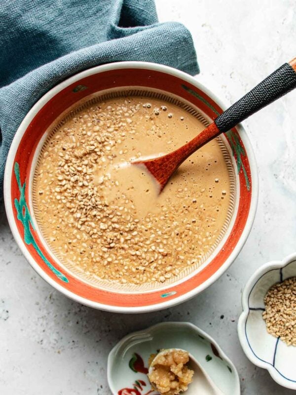 A bowl of creamy, light brown miso sesame dressing topped with sesame seeds, with a wooden spoon resting inside. A blue cloth is nearby, and small dishes with extra sesame seeds and miso paste sit beside the bowl.