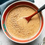A bowl filled with creamy miso sesame dressing, topped with sesame seeds and being stirred with a wooden spoon. The bowl features a decorative red and teal pattern along the inside edge.