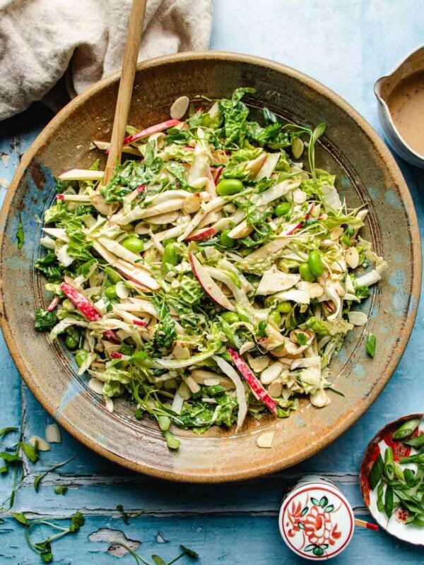 A large bowl of colorful Napa cabbage salad with shredded cabbage, sliced apples, almonds, green edamame, and herbs mixed with dressing. A wooden spoon rests in the bowl on a blue surface, with dressing and napkin nearby.