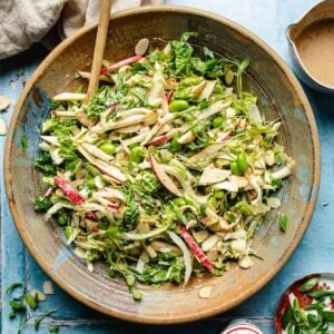 A large bowl of Napa cabbage salad with sliced apples, edamame, greens, and slivered almonds. A wooden spoon rests inside the bowl; a cup of brown dressing sits nearby on a blue surface.