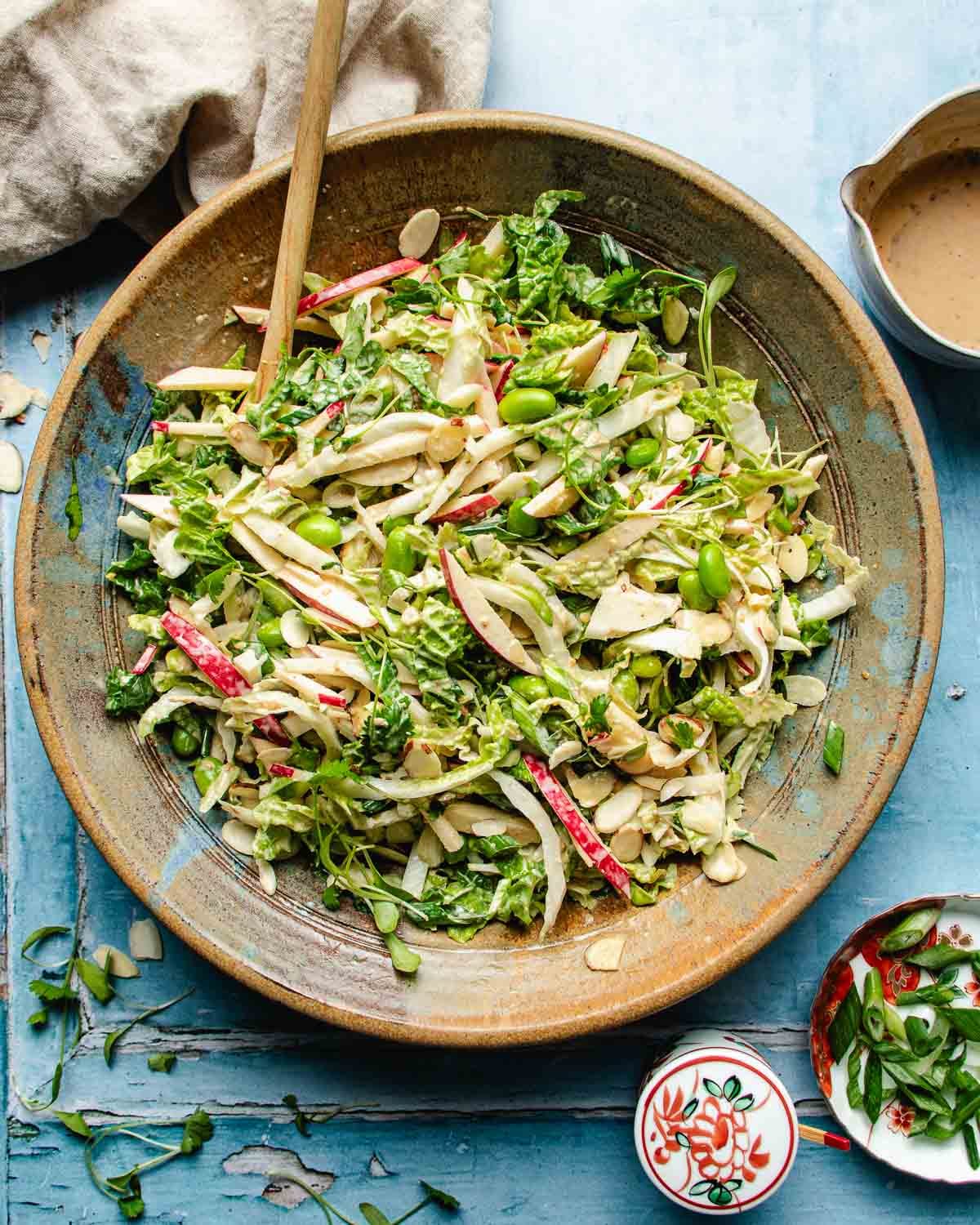 A large bowl of colorful Napa cabbage salad with shredded cabbage, sliced apples, almonds, green edamame, and herbs mixed with dressing. A wooden spoon rests in the bowl on a blue surface, with dressing and napkin nearby.