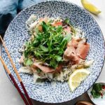 A patterned blue and white bowl filled with daikon ribbons, prosciutto, green leafy sprouts, black sesame seeds, and a bed of shaved daikon salad. Lemon wedges and chopsticks sit beside the bowl, with a blue napkin and small dish nearby.