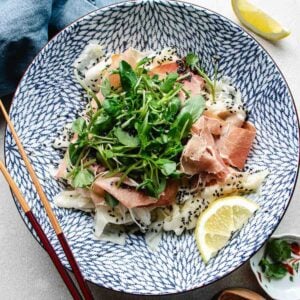 A patterned blue and white bowl filled with daikon ribbons, prosciutto, green leafy sprouts, black sesame seeds, and a bed of shaved daikon salad. Lemon wedges and chopsticks sit beside the bowl, with a blue napkin and small dish nearby.