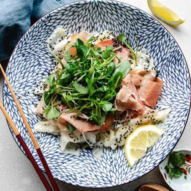 A patterned blue and white bowl filled with daikon ribbons, prosciutto, green leafy sprouts, black sesame seeds, and a bed of shaved daikon salad. Lemon wedges and chopsticks sit beside the bowl, with a blue napkin and small dish nearby.