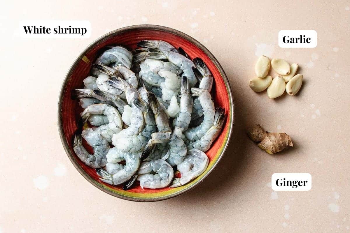 A bowl of raw white shrimp for Thai cucumber shrimp salad is placed on a light surface. To the right, several garlic cloves and a piece of ginger are arranged. Each ingredient is labeled: White shrimp, Garlic, and Ginger.