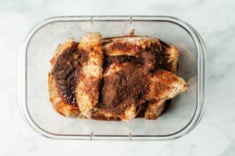 Raw chicken wings in a clear container, coated with a generous amount of dark, dry spice rub inspired by Asian chicken marinades, on a white marble surface.