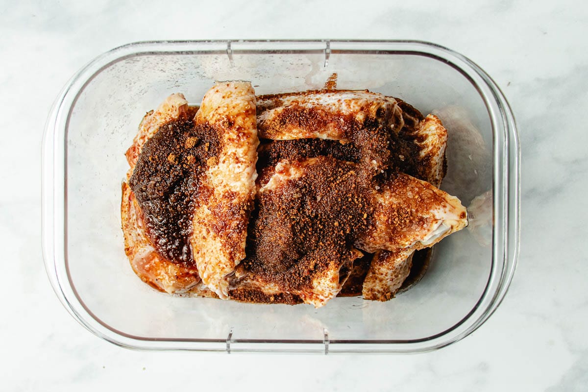 Raw chicken wings in a clear container, coated with a generous amount of dark, dry spice rub inspired by Asian chicken marinades, on a white marble surface.