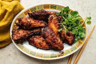 A plate of crispy, glazed chicken wings infused with Asian chicken marinades is garnished with fresh green herbs and sits on a decorative dish next to a yellow napkin and wooden chopsticks.