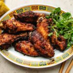 A decorative oval plate with a Chinese pattern border holds several browned, glazed chicken marinated with Asian chicken marinades and garnished with fresh green herbs, placed on a light-colored surface.
