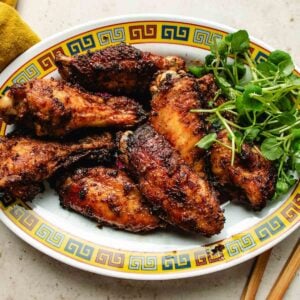 A decorative oval plate with a Chinese pattern border holds several browned, glazed chicken marinated with Asian chicken marinades and garnished with fresh green herbs, placed on a light-colored surface.