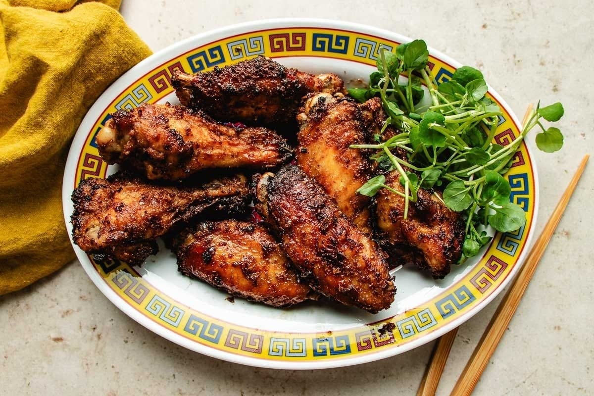 A plate of crispy, glazed chicken wings infused with Asian chicken marinades is garnished with fresh green herbs and sits on a decorative dish next to a yellow napkin and wooden chopsticks.
