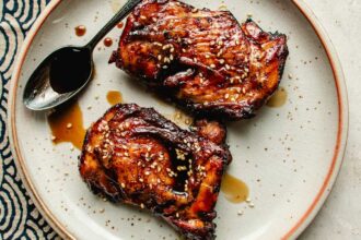 Two pieces of glazed, grilled chicken featuring Asian chicken marinades are topped with sesame seeds on a white plate with a brown rim; a spoon with extra sauce rests beside the chicken.