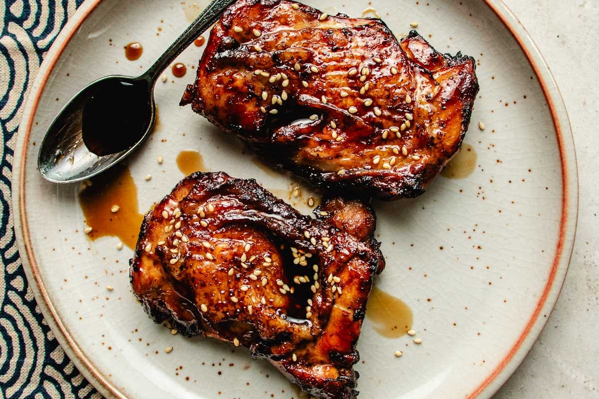 Two pieces of glazed, grilled chicken featuring Asian chicken marinades are topped with sesame seeds on a white plate with a brown rim; a spoon with extra sauce rests beside the chicken.