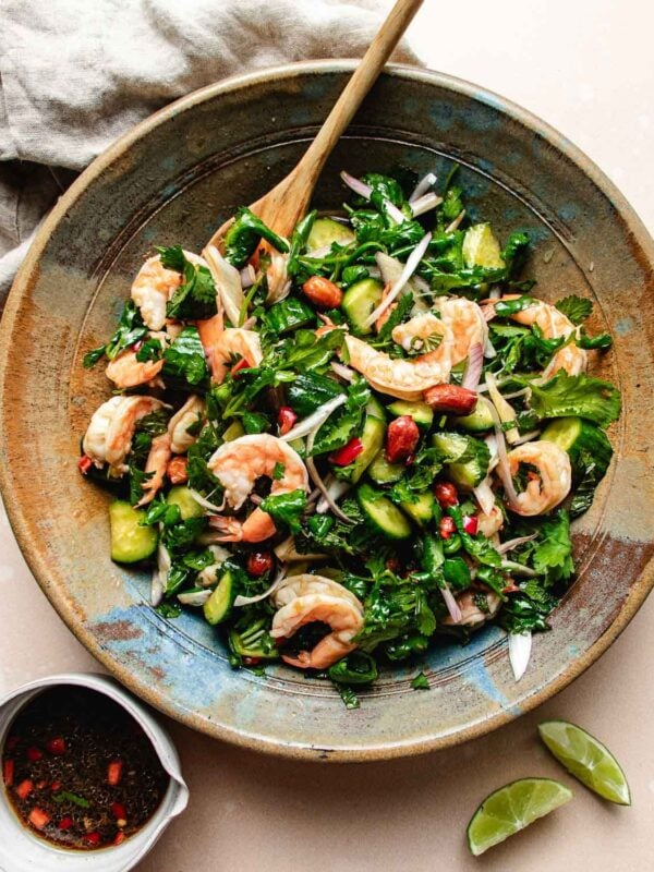 A large bowl of Thai cucumber shrimp salad with fresh herbs, onions, and peanuts, mixed with a wooden spoon. Lime wedges and a small bowl of chili-flake dressing sit nearby on the table.