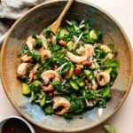 A rustic bowl filled with Thai cucumber shrimp salad featuring shrimp, cucumber slices, red onion, fresh herbs, roasted peanuts, and red chili slices, with a wooden spoon and a lime wedge nearby.