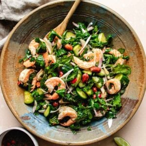 A rustic bowl filled with Thai cucumber shrimp salad featuring shrimp, cucumber slices, red onion, fresh herbs, roasted peanuts, and red chili slices, with a wooden spoon and a lime wedge nearby.