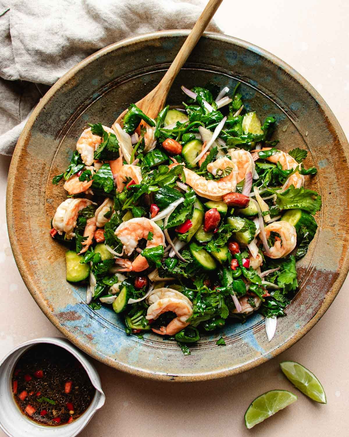 A large bowl of Thai cucumber shrimp salad with fresh herbs, onions, and peanuts, mixed with a wooden spoon. Lime wedges and a small bowl of chili-flake dressing sit nearby on the table.