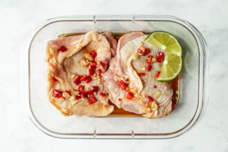 Two raw chicken thighs marinate in a rectangular glass container with chopped red chili, garlic, sauce, and two lime slices on top—perfect for those seeking bold Asian chicken marinades. The container rests on a white surface.