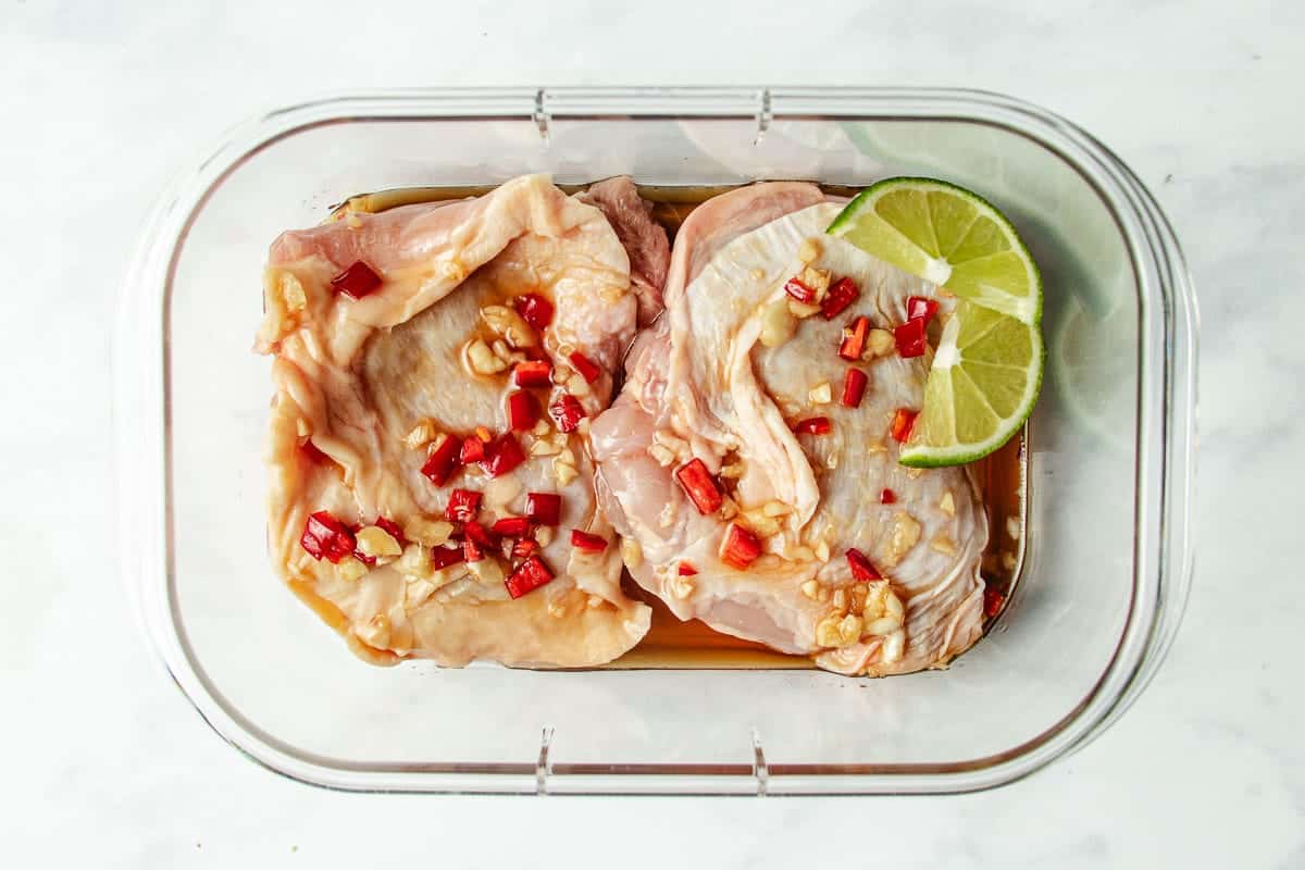 Two raw chicken thighs marinate in a rectangular glass container with chopped red chili, garlic, sauce, and two lime slices on top—perfect for those seeking bold Asian chicken marinades. The container rests on a white surface.