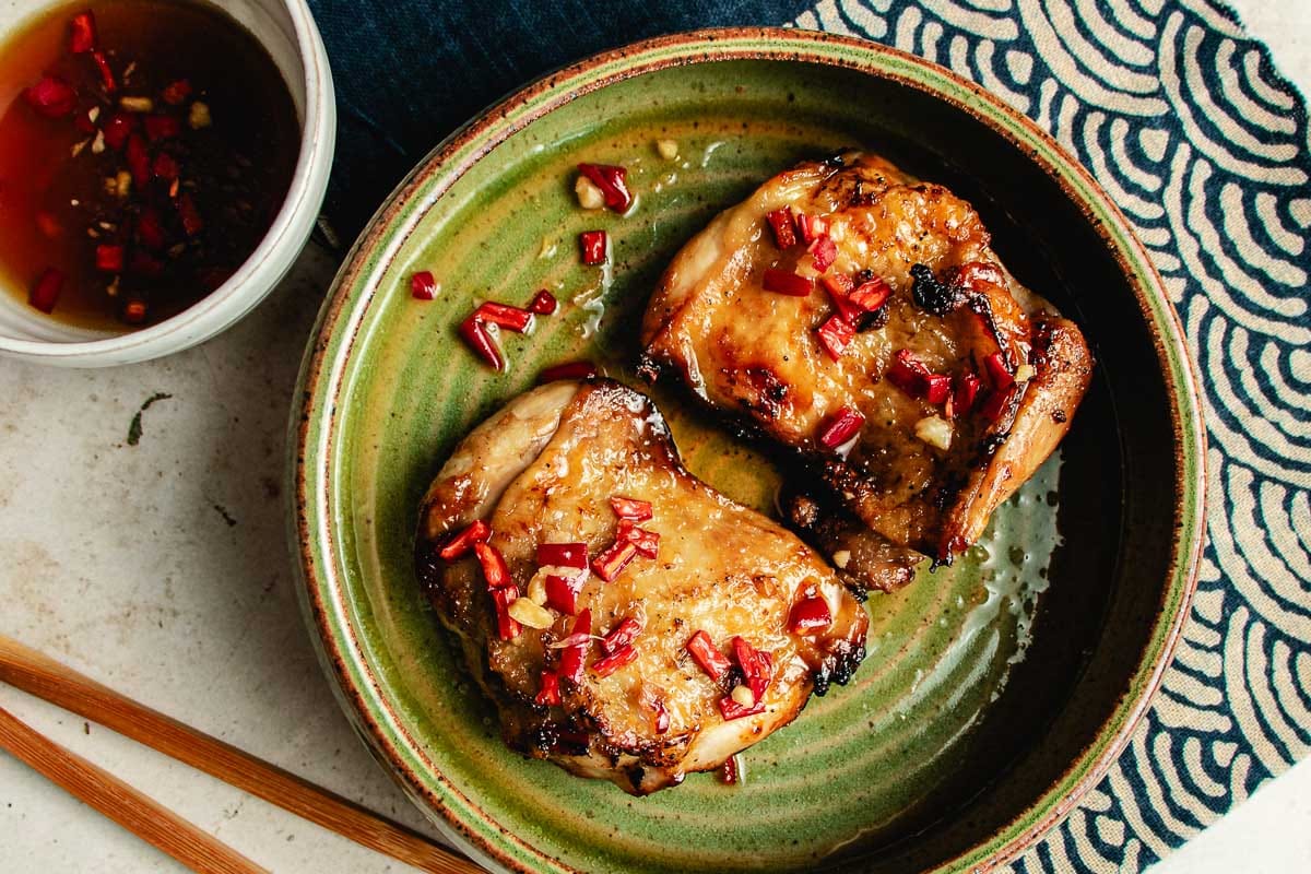 Two pieces of grilled chicken, marinated in savory Asian chicken marinades, are topped with chopped red chilies on a green ceramic plate, accompanied by a small bowl of sauce and wooden chopsticks on a patterned cloth.