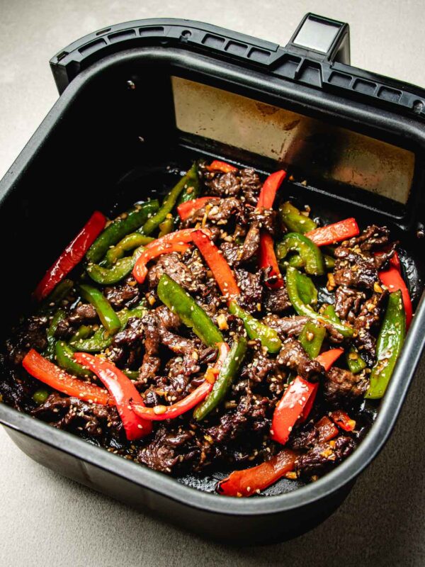 Air fryer beef stir fry featuring sliced beef with red and green bell peppers and sesame seeds in an air fryer basket, cooked and ready to serve.