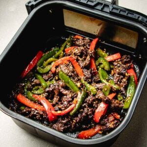 Air fryer beef stir fry features tender beef strips, vibrant red and green bell peppers, and a sprinkle of sesame seeds, all cooked together in an air fryer basket to create a colorful and flavorful stir-fry dish.