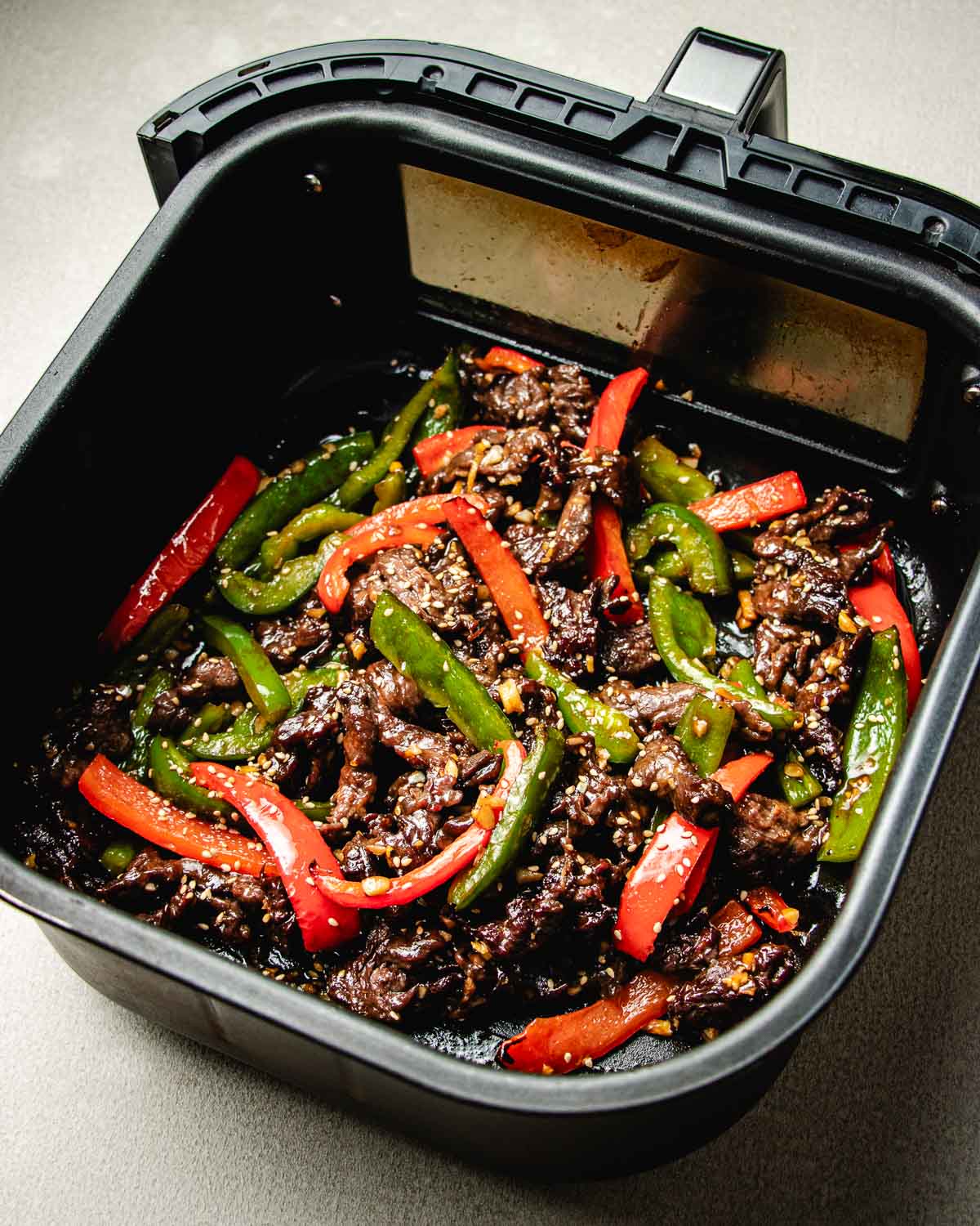 Air fryer beef stir fry featuring sliced beef with red and green bell peppers and sesame seeds in an air fryer basket, cooked and ready to serve.