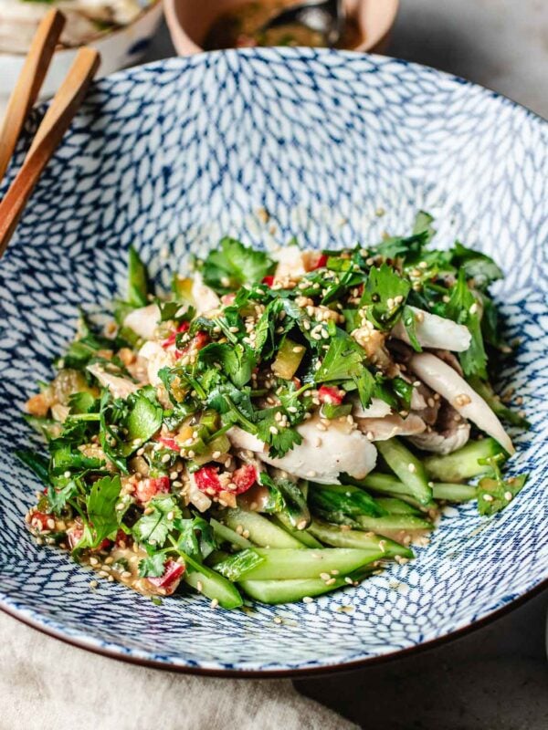A patterned bowl filled with a Bang Bang Chicken Taiwanese Style salad made of shredded chicken, sliced cucumber, cilantro, red chilies, and sesame seeds. Wooden chopsticks rest on the bowl’s edge.