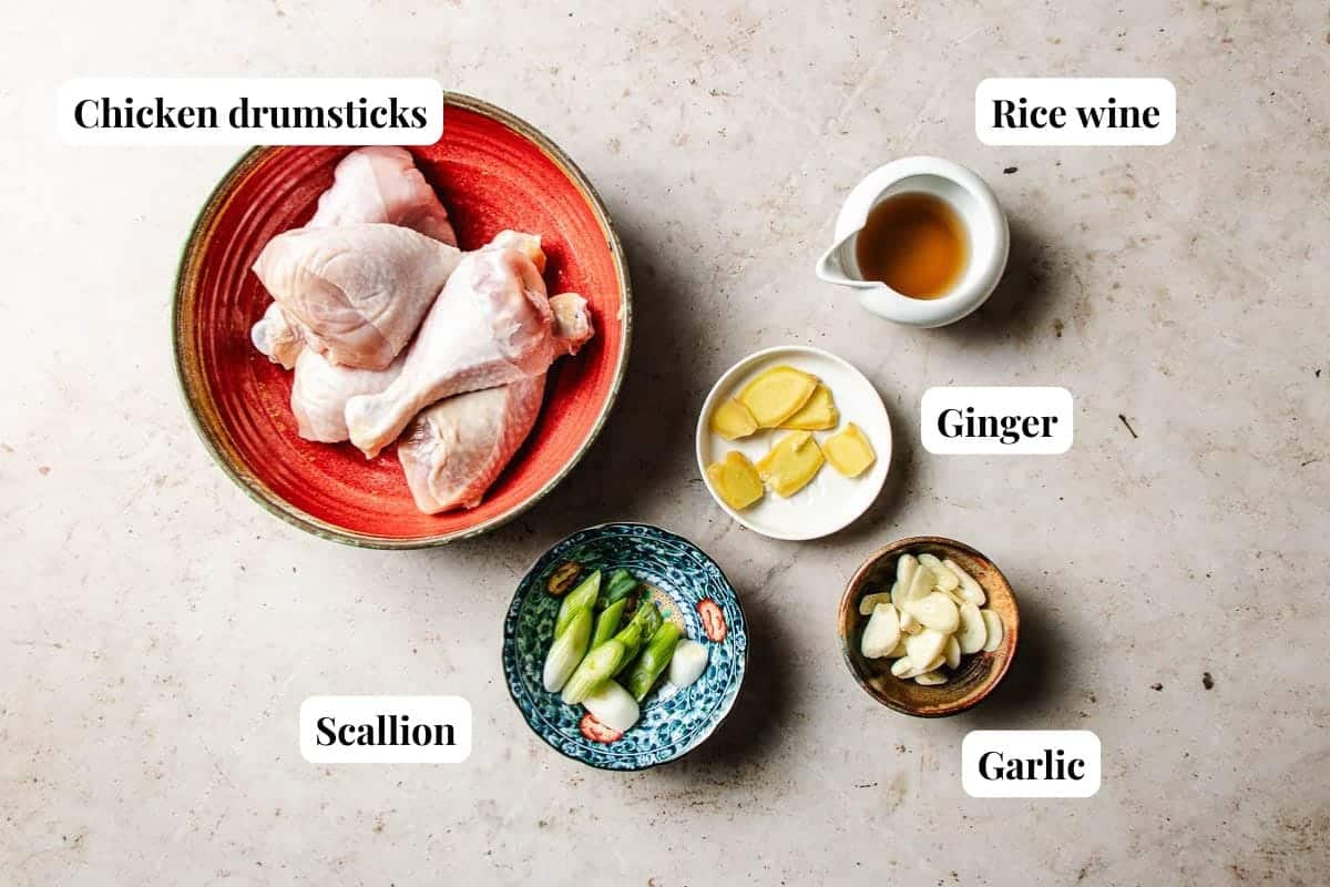 Chicken drumsticks in a red bowl, rice wine in a white pitcher, sliced ginger, scallion pieces in a blue bowl, and sliced garlic in a small brown bowl, all labeled on a light surface—perfect ingredients for preparing Bang Bang Chicken Taiwanese Style.